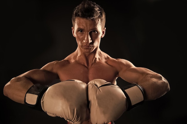 Macho with muscular chest posing with boxing gloves black background Sportsman in boxing gloves shows his muscular arms Athlete on confident face demonstrates muscular body Boxing concept