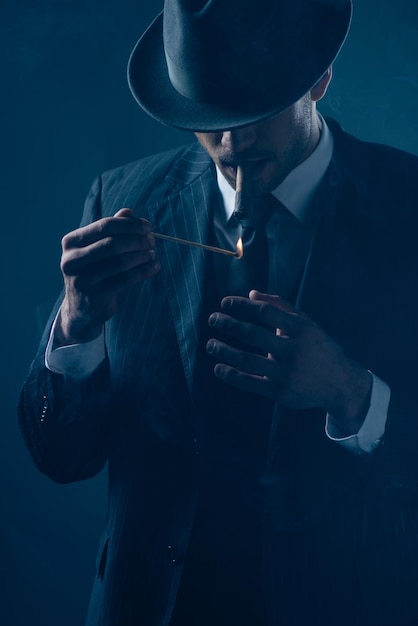 Mafioso with covered eyes with felt hat lighting cigar on dark blue