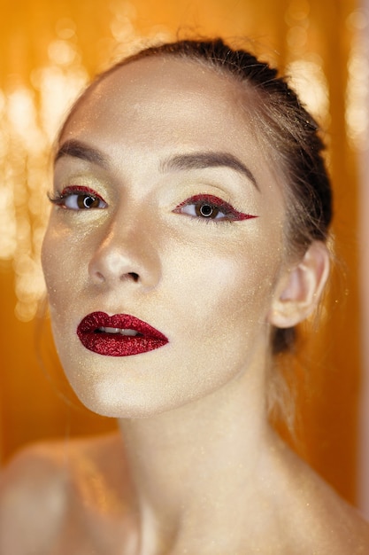 Magic Girl Portrait in Gold. Golden Makeup, close-up portrait in studio shot, color. Beauty Model Girl with perfect bright make-up, red lips, golden maroon jewellery. Sexy lady makeup Holiday party.