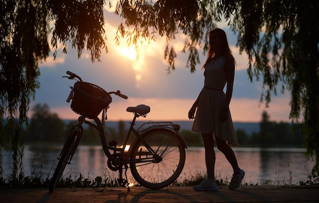 Magical sunset landscape Silhouette of woman in dress and retro bicycle with basket against lake