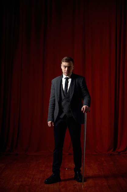 Magician wearing suit standing with cane on stage decorated red velvet curtain