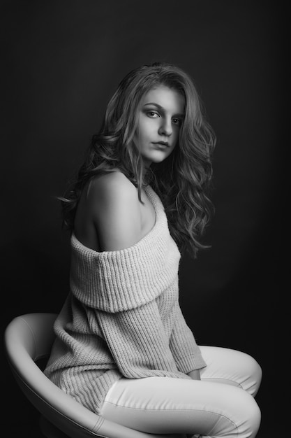 Magnificent brunette woman with long curly hair wears knitted sweater with naked shoulders posing in the shadow. Monochrome shot