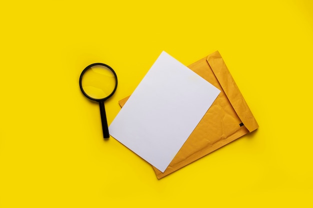 Magnifying glass, blank white sheet and envelope on a yellow surface.