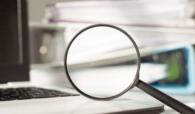 Magnifying glass on a laptop amid a stack of office folders with documents. Business and Search Concept.