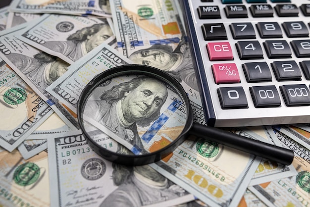 Magnifying glass on pile of US dollar money banknotes with calculator isolated on wooden table