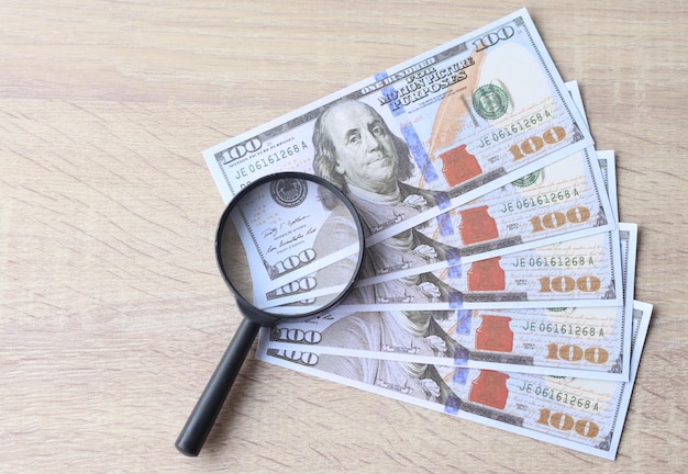 Magnifying glass with fan of 100 dollar bills on wooden table