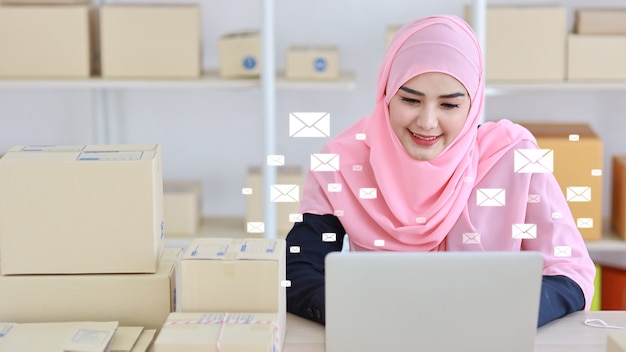 Mail communication technology concept. Young smart asian muslim businesswoman working on computer, mobile smartphone receiving and sending customers e-mail with notification message icons above device