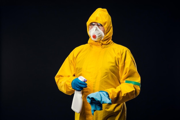 A maintenance worker in a protective yellow suit and protective mask holds a disinfectant spray on a black background