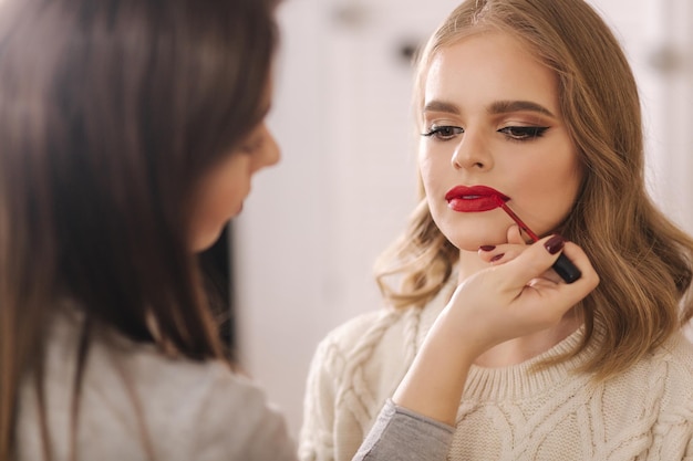 Makeup artist applies red lipstick Hand of makeup master painting lips of young beauty blond hair model Make up in process Closeup view