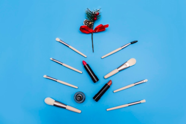 Makeup brushes are laid out in the shape of a fir tree on a blue background along with red lipsticks and eye shadow