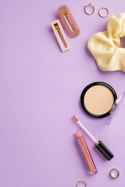 Makeup concept Top view vertical photo of scrunchy lip gloss compact powder gold rings and two hairpins on isolated lilac background