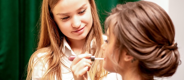 Makeup in the process The makeup artist applies pink gloss lipstick on the lips of the beautiful face of the young caucasian woman in a beauty salon