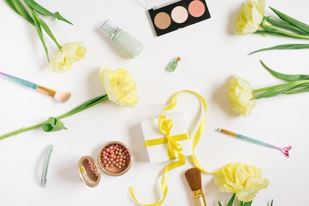 Makeup products with yellow tulip flowers and a gift box with a bow on a white background