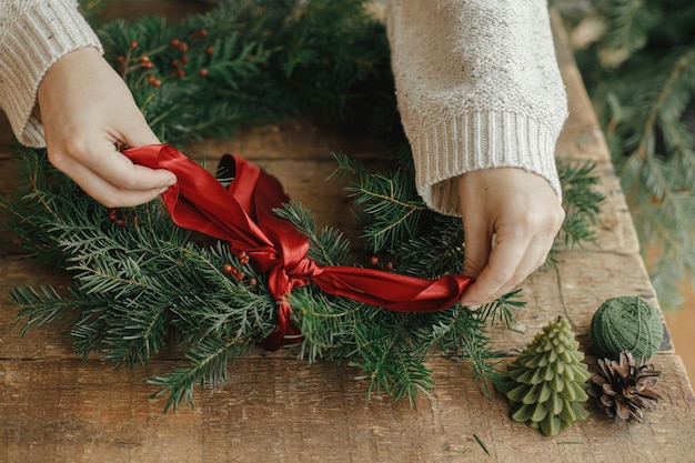 Making Christmas wreath Woman hands decorating christmas wreath with red ribbon on rustic table