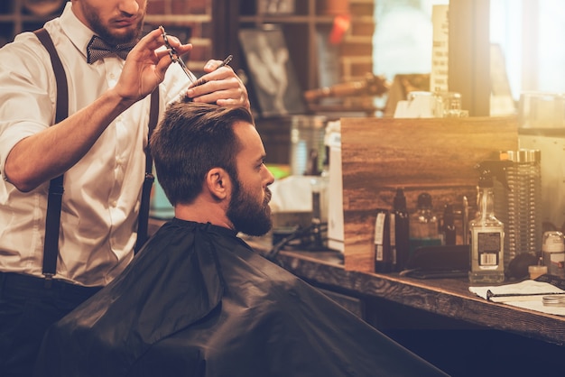 Making hair look magical. Young bearded man getting haircut by hairdresser while sitting in chair at barbershop