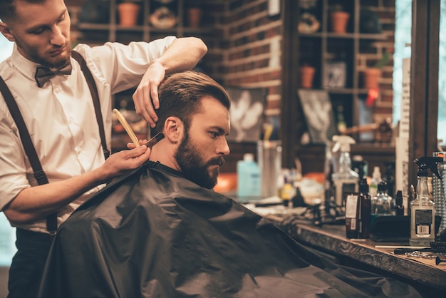 Making hair look magical. Young bearded man getting haircut with straight edge razor by hairdresser 