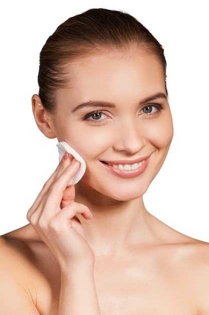 Making her face clean and fresh. Beautiful young shirtless woman wiping her face with sponge and smiling while standing isolated on white background