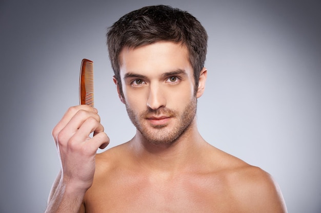 Making his unique style. Handsome young shirtless man holding a comb and looking at camera while standing against grey background