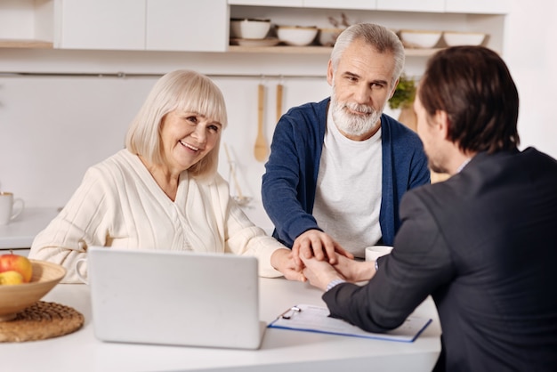 Making success deal . Smiling satisfied old couple sitting at home and concluding agreement with real estate agent while shaking hands and expressing happiness