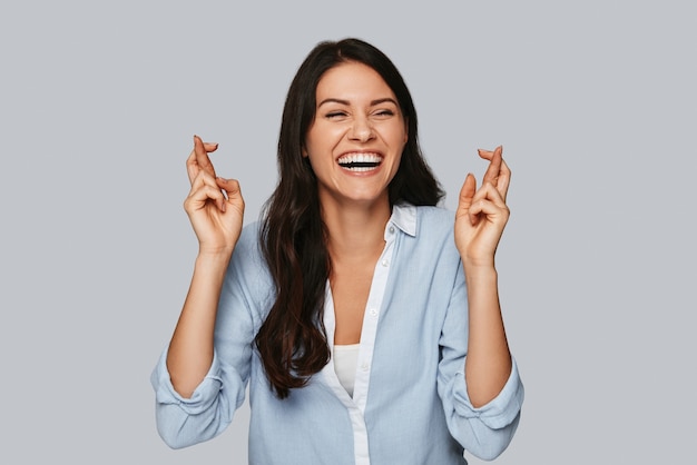 Making a wish. Attractive young woman crossing fingers and smiling while standing against grey background