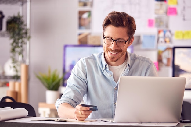 Male Architect In Office Working At Desk Making Online Purchase Using Credit Card On Laptop