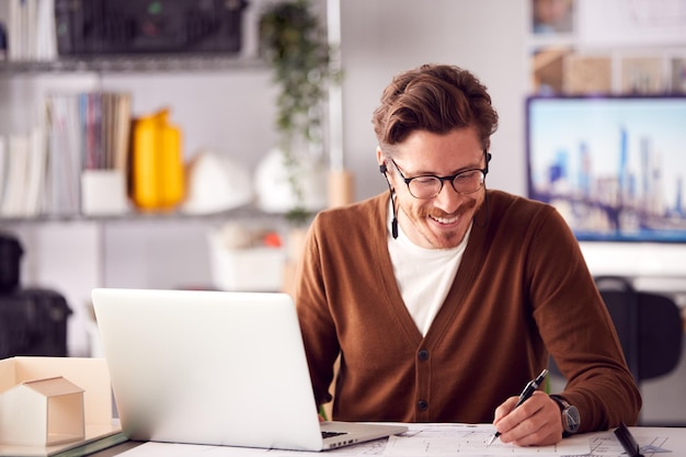 Male Architect In Office Working On Plan At Desk Taking Phone Call On Wireless Earphones