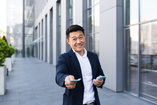 Male asian businessman rejoices holding a lot of cash looking at the camera giving money