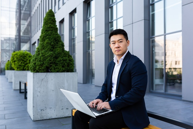 Male asian businessman working on laptop online banking on computer near office center looking at camera