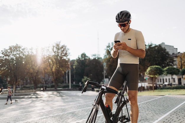 Male athlete in helmet and glasses sitting on black bike with modern smartphone in hands Caucasian cyclist texting on mobile while taking break during workout