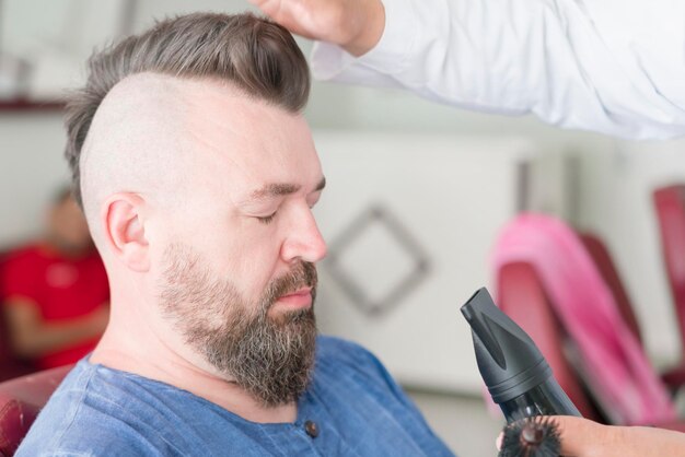 Male barber makes hair styling using a hair dryer of a adult bearded man with a mohawk