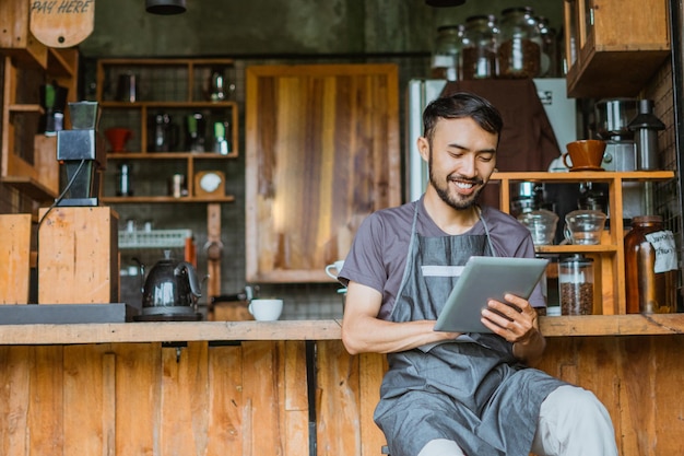 Male barista in apron using the digital tablet while sitting