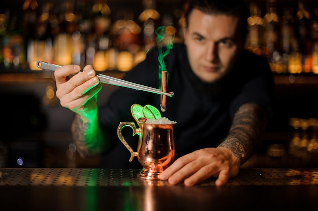 Male bartender adding to a cocktail in the cooper glass with a dried orange aromatic smoked cinnamon with tweezers on the bar counter