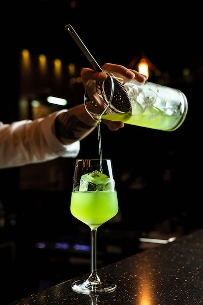 Male bartender prepairing a cocktail, pouring a green drink from a mixing glass through a strainer into a wine glass with an ice cube