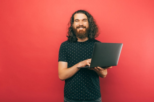 Male bearded hipster is holding a laptop and smiling at the camera in a studio