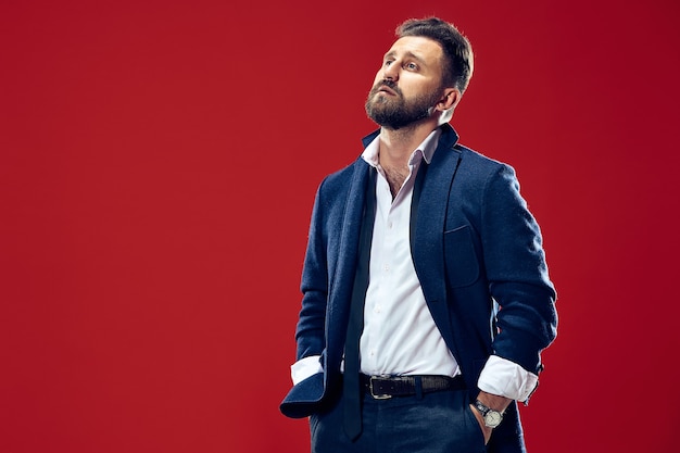 Male beauty concept portrait of a fashionable young man with stylish haircut wearing trendy suit posing over red wall