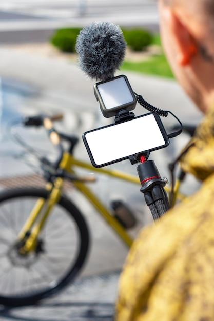 A male blogger holds a tripod in his hands with a smartphone mockup with a white screen and a light with a microphone on the background of a bicycle