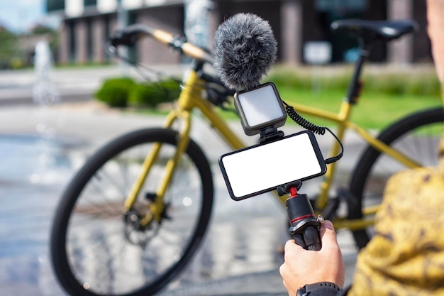 A male blogger holds a tripod in his hands with a smartphone mockup with a white screen and a light with a microphone on the background of a bicycle