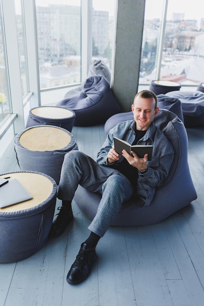 A male businessman in a casual suit resting in a comfortable pouf while working an attractive freelancer in a suit works remotely and checks notes in a notebook