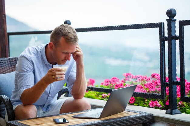 Male businessman working on laptop at rooftop cafe with beautiful panoramic view. Guy blogger drinks coffee and works on a computer while traveling