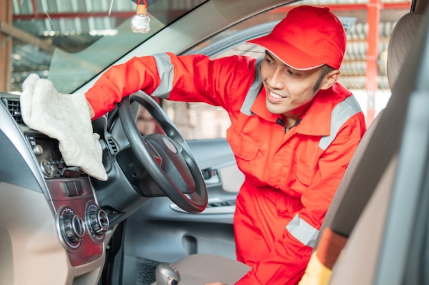 Male car cleaner wearing red uniform is wiping car interior dashboard in car salon