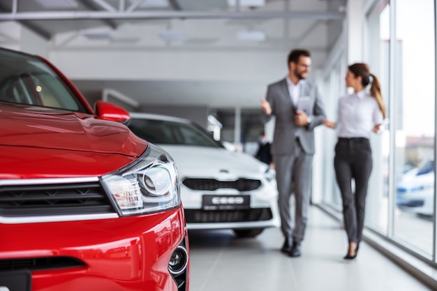 Male car seller in suit walking around car salon with woman who wants to buy a car and talking about specifications of cars