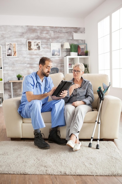 Male care taker and senior woman in nursing home on couch using tablet computer.