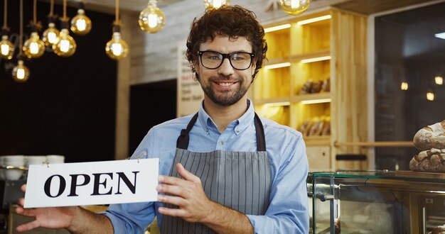 Male caucasian smiled bakery vendor posing with a signboard OPEN and smiling in the shop.