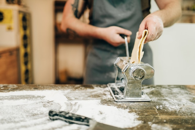 Male chef in apron works with pasta machine on wooden kitchen table. Homemade spaghetti cooking process