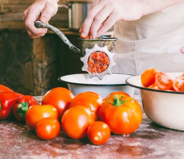 Male chef grinds pieces of ripe tomatoes in an old vintage manual grinder to make homemade sauce