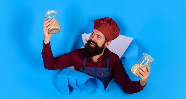 Male chef in hat and apron holds jars with cereals looking through paper hole breakfast and healthy