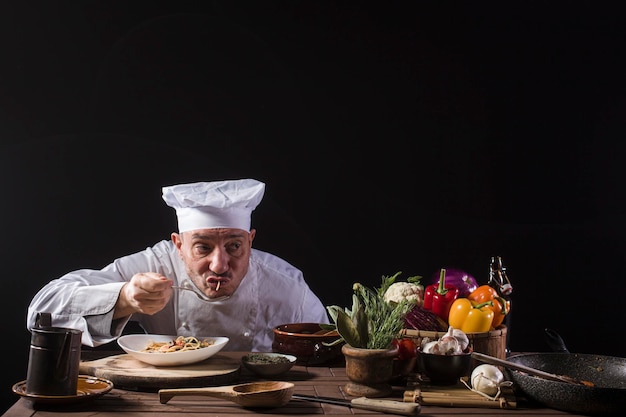 Male chef in a restaurant kitchen wearing white uniform tasting spaghetti with fresh vegetables