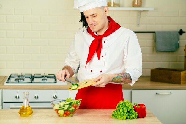 Male chef with dreadlocks adding fresh cucumber slices in glass bowl with salad in kitchen A man cooks Vitamins dietary and sports nutrition Vegetarian food