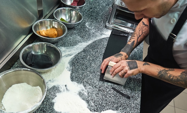 male chef with tattoos on his hands cutting dough for homemade pasta on kitchen table