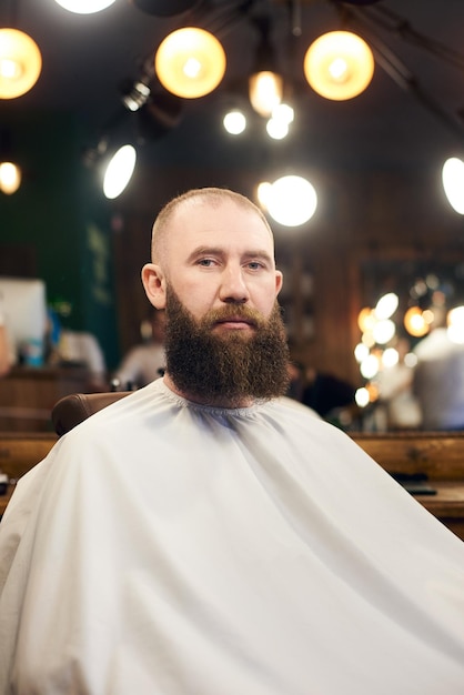 Male client with beard sitting in hairdresser chair Serious man with long brown beard Modern popular lumberjack style